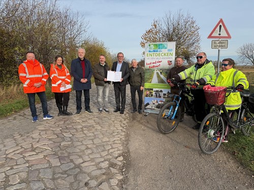 Zum Bild (v.l.): Fachdienstleiter Tilo Wechselberger, Jana Elbe, Ingenieur Lutz Koslowski, Verbandsgemeinde-Bürgermeister Michael Stöhr, Landrat Markus Bauer, Egelns Bürgermeister Reinhard Luckner und Hakeborns stellvertretender Bürgermeister Dirk Bartsch sowie zwei Radfahrer auf der Kreisstraße, die bis 2027 zu einer Fahrradstraße umgebaut werden soll. Bildquelle: Marko Jeschor / Pressestelle Salzlandkreis